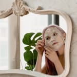 A woman looking at her reflection while applying a facial sheet mask at home.