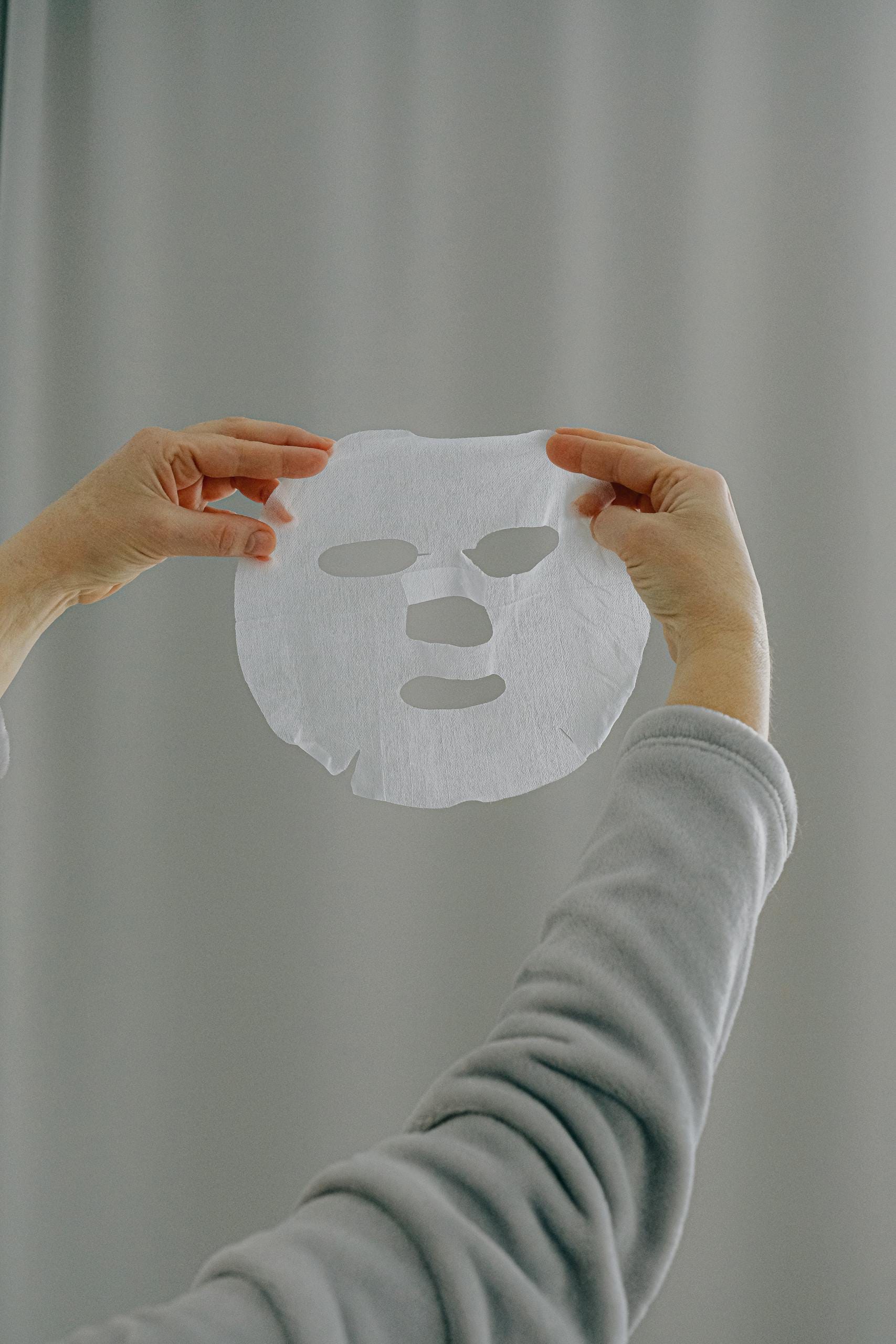 Hands presenting a white sheet mask against a neutral background, promoting skincare and wellness.