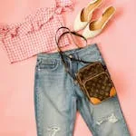 A trendy flat lay of a chic summer outfit featuring denim jeans, crop top, heels, and a stylish bag on a pink background.