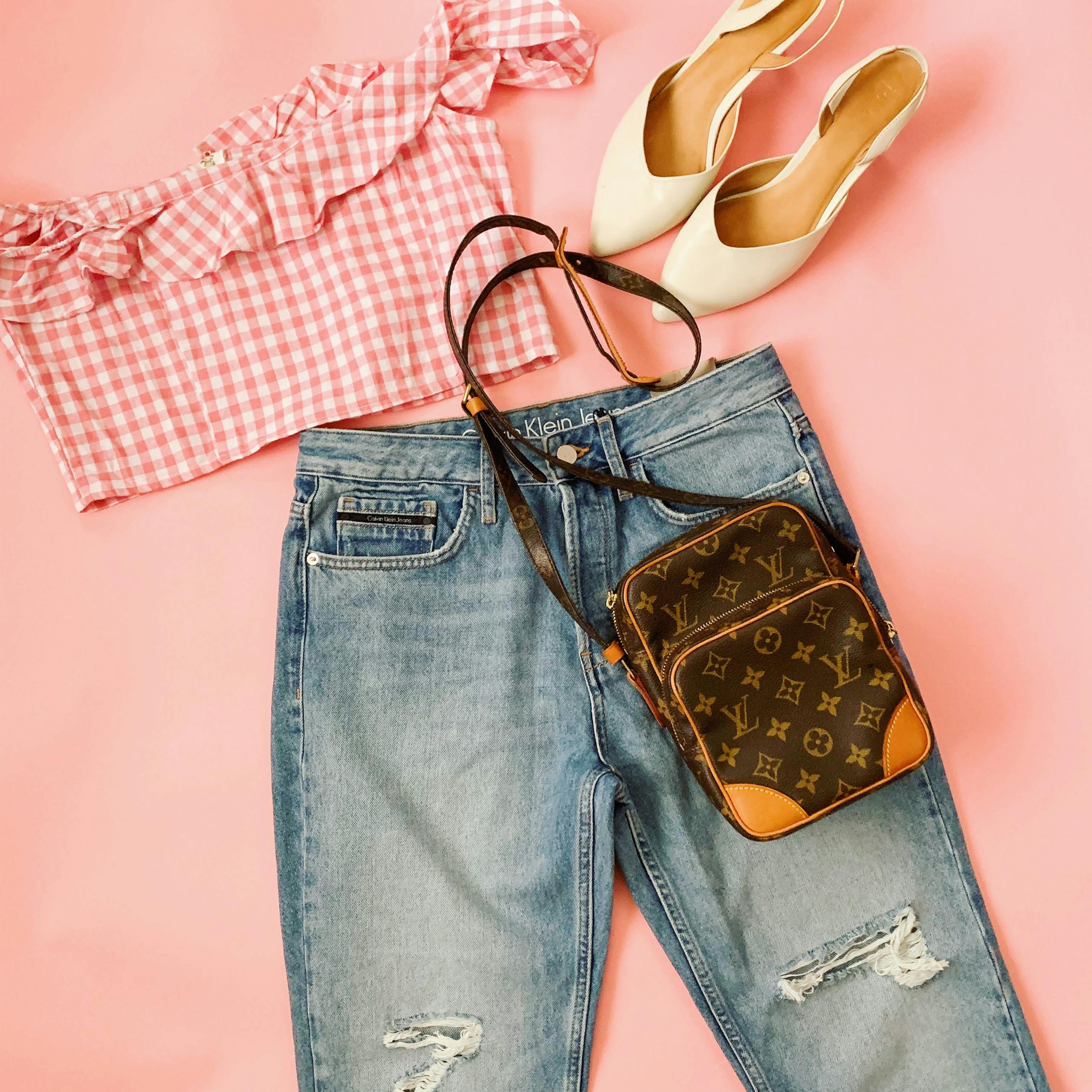 A trendy flat lay of a chic summer outfit featuring denim jeans, crop top, heels, and a stylish bag on a pink background.