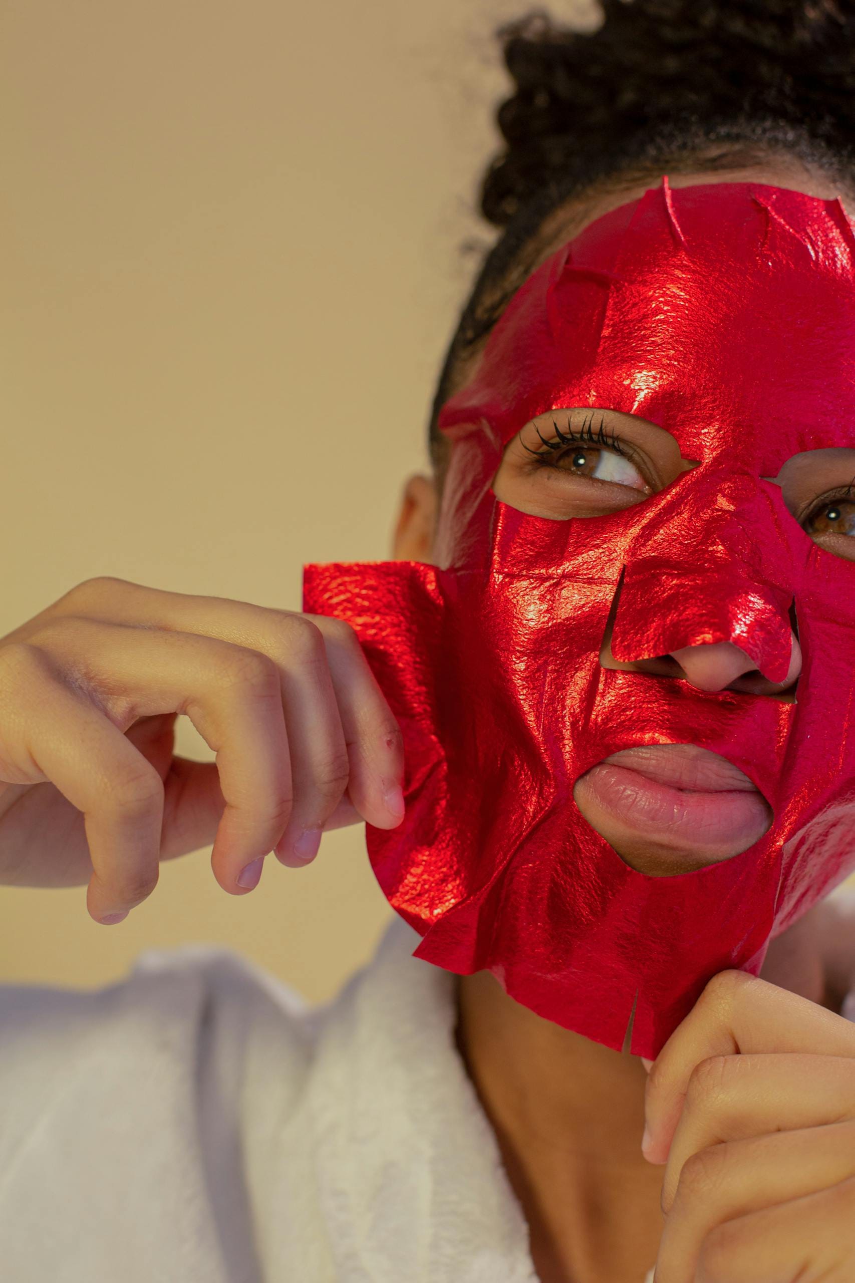 A young woman in a bathrobe removing a red facial mask, focusing on skincare and wellness.