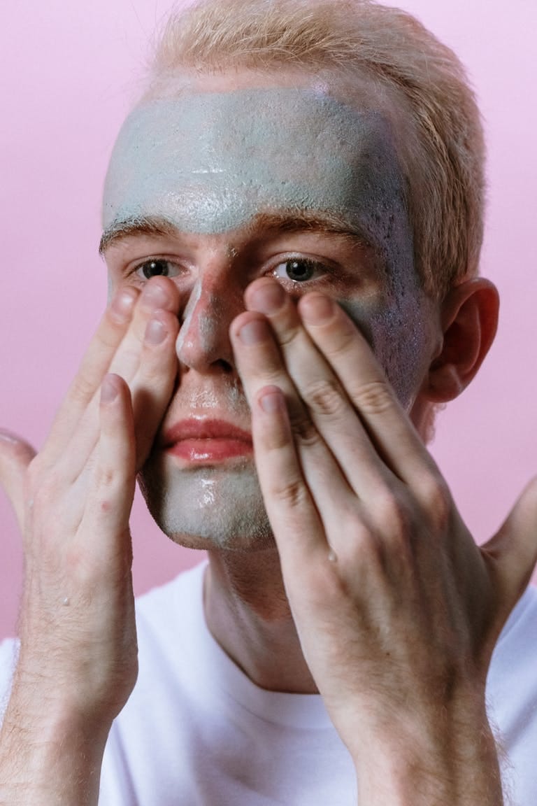 Young man applying face mask as part of a skincare routine with a pink background.