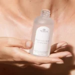 Woman examining her skin in a modern bathroom mirror with a minimal selection of skincare products beside her