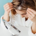 Woman in bathrobe checking grey hair roots in front of a mirror indoors.